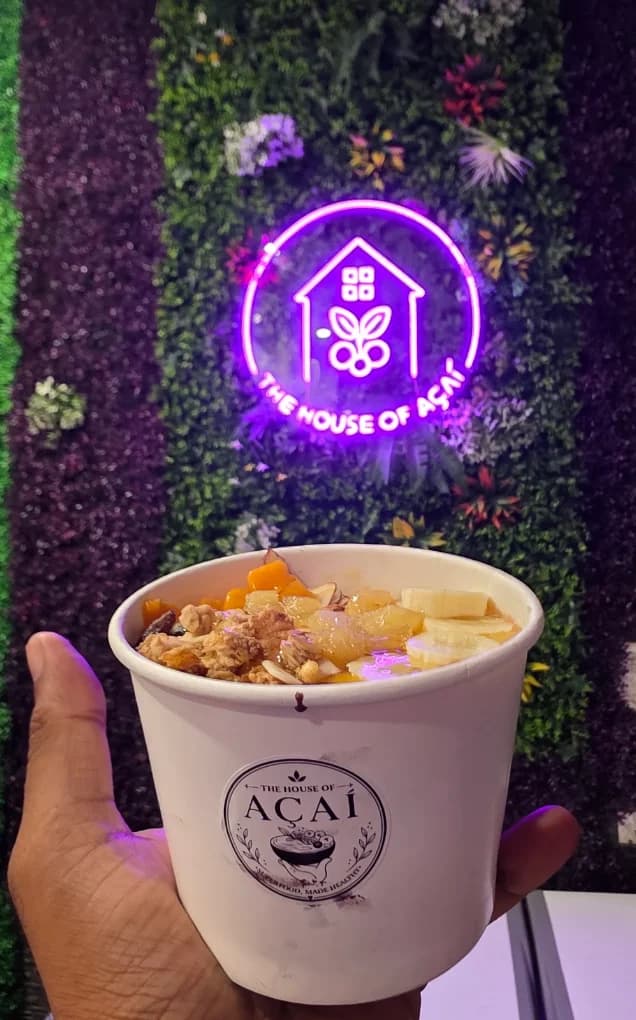 A tropical açaí bowl held up beneath the glowing neon House of Açaí sign on the café's moss wall